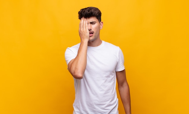 Hombre guapo joven que parece somnoliento, aburrido y bostezando, con dolor de cabeza y una mano cubriendo la mitad de la cara