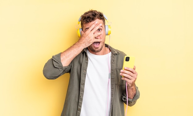 Hombre guapo joven que parece sorprendido, asustado o aterrorizado, cubriendo la cara con auriculares de mano y concepto de teléfono inteligente