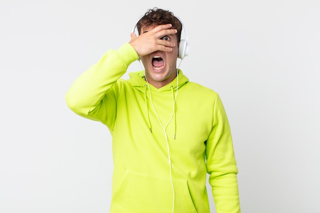 Hombre guapo joven que parece sorprendido, asustado o aterrorizado, cubriendo la cara con la mano y los auriculares