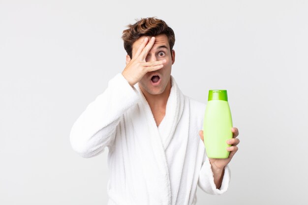 Hombre guapo joven que parece sorprendido, asustado o aterrorizado, cubriendo la cara con la mano con una bata de baño y sosteniendo una botella de champú