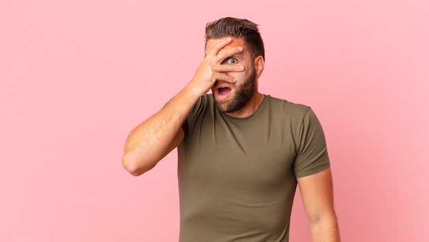 Hombre guapo joven que parece sorprendido, asustado o aterrorizado, cubriendo la cara con la mano