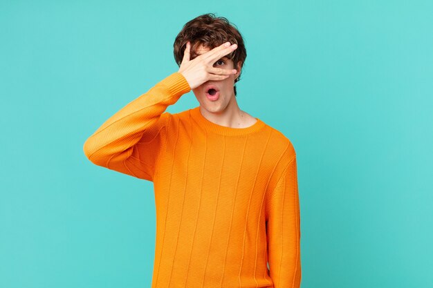 Hombre guapo joven que parece sorprendido, asustado o aterrorizado, cubriendo la cara con la mano