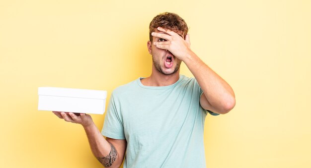 Hombre guapo joven que parece sorprendido, asustado o aterrorizado, cubriendo el rostro con la mano. concepto de caja blanca