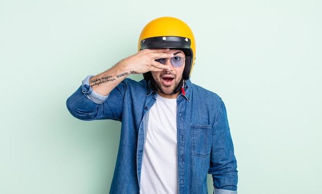 Hombre guapo joven que parece sorprendido, asustado o aterrorizado, cubriendo el rostro con la mano. concepto de casco de moto