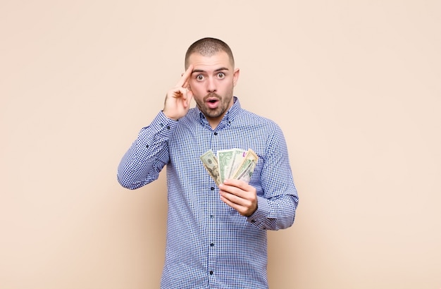 Hombre guapo joven que parece sorprendido, con la boca abierta, conmocionado, dándose cuenta de un nuevo pensamiento, idea o concepto con billetes de dólar