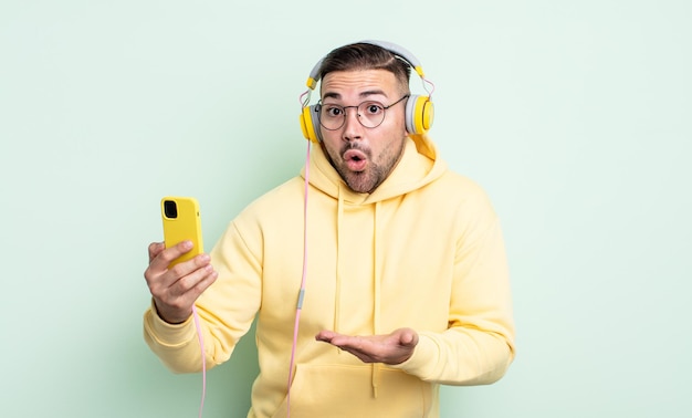 Hombre guapo joven que parece sorprendido y conmocionado, con la mandíbula caída sosteniendo un objeto. auriculares y concepto de teléfono inteligente