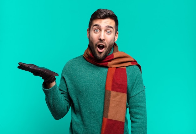 Hombre guapo joven que parece sorprendido y conmocionado, con la mandíbula caída sosteniendo un objeto con una mano abierta en el costado. concetp frío e invierno