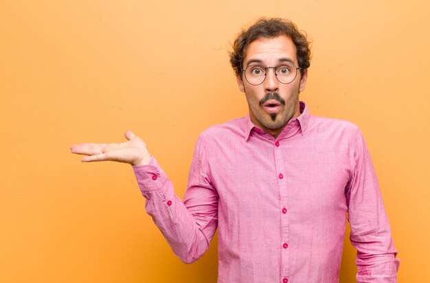 Hombre guapo joven que parece sorprendido y conmocionado, con la mandíbula caída sosteniendo un objeto con la mano abierta en el lado contra la pared naranja