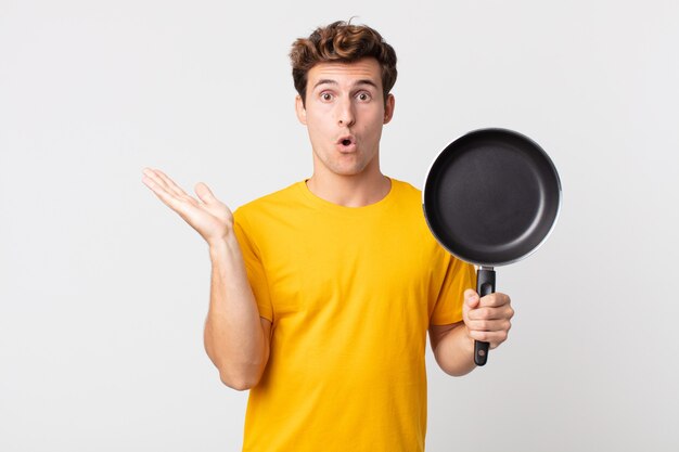 Hombre guapo joven que parece sorprendido y conmocionado, con la mandíbula caída sosteniendo un objeto y sosteniendo una sartén