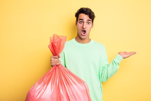 Hombre guapo joven que parece sorprendido y consternado, con la mandíbula caída sosteniendo un objeto. concepto de bolsa de basura