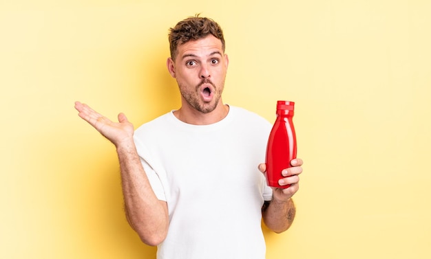 Hombre guapo joven que parece sorprendido y consternado, con la mandíbula caída sosteniendo un objeto. concepto de salsa de tomate