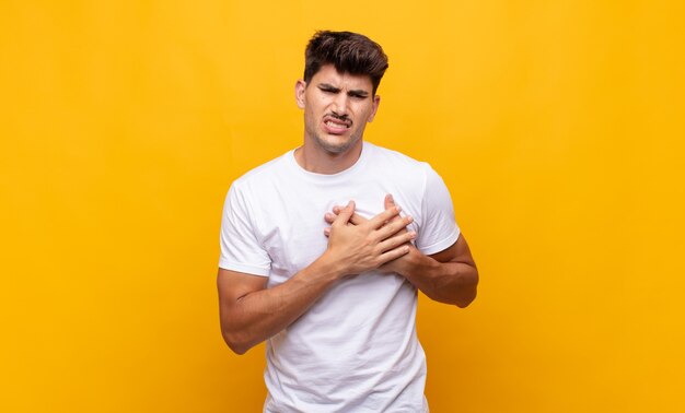 Hombre guapo joven que parece triste, herido y con el corazón roto, sosteniendo ambas manos cerca del corazón, llorando y sintiéndose deprimido