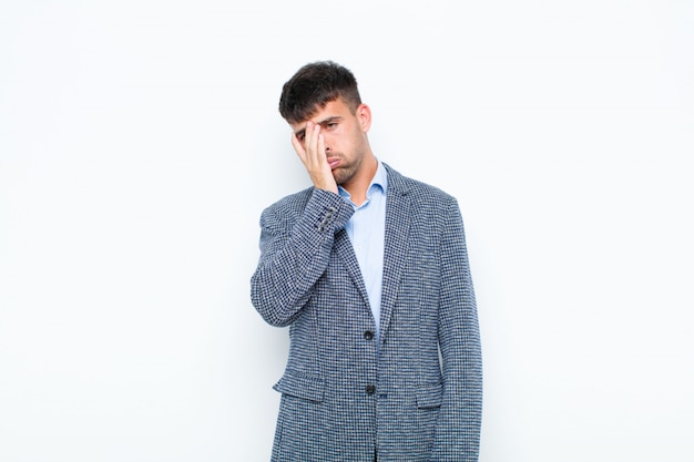 Hombre guapo joven que se siente aburrido, frustrado y con sueño después de una tarea tediosa, aburrida y tediosa, sosteniendo la cara con la mano contra la pared blanca