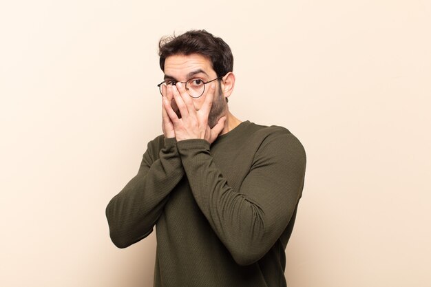 Hombre guapo joven que se siente asustado o avergonzado, mirando o espiando con los ojos medio cubiertos con las manos