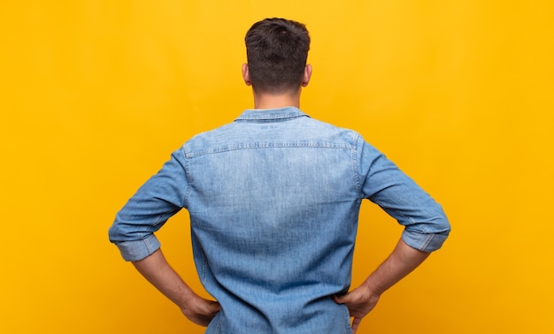 Hombre guapo joven que se siente confundido o lleno o con dudas y preguntas, preguntándose, con las manos en las caderas, vista trasera