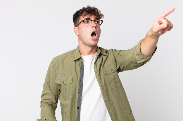 Hombre guapo joven que se siente conmocionado y sorprendido, apuntando y mirando hacia arriba con asombro con una mirada asombrada y con la boca abierta.