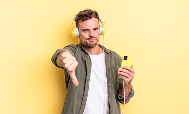 Hombre guapo joven que se siente cruzado, mostrando los pulgares hacia abajo en los auriculares y el concepto de teléfono inteligente