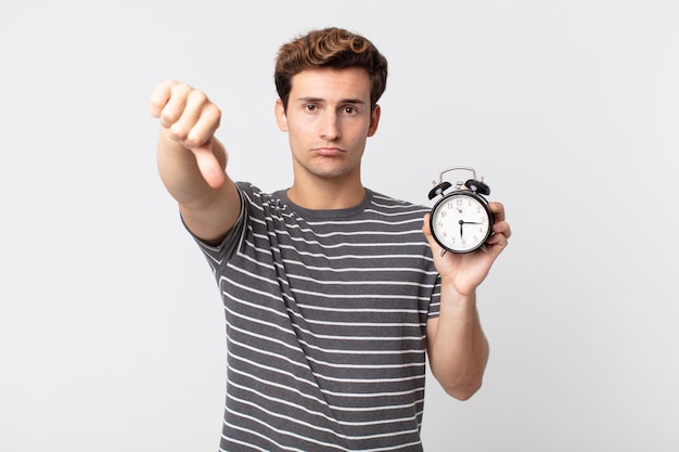 Hombre guapo joven que se siente cruzado, mostrando los pulgares hacia abajo y sosteniendo un reloj despertador