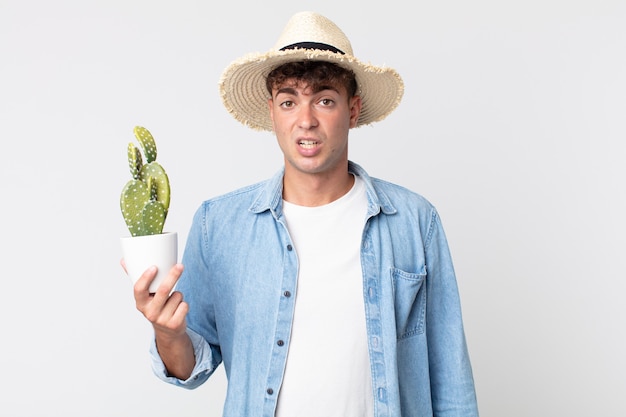 Hombre guapo joven que se siente desconcertado y confundido. granjero sosteniendo un cactus decorativo