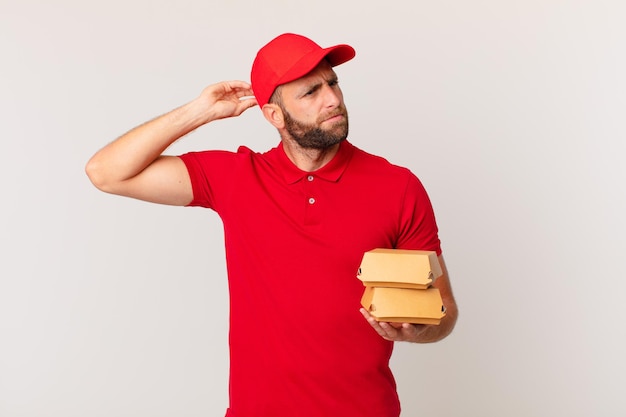 Hombre guapo joven que se siente desconcertado y confundido, rascándose la cabeza entregando hamburguesa concepto