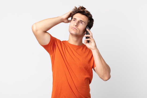 Hombre guapo joven que se siente desconcertado y confundido, rascándose la cabeza y hablando con un teléfono inteligente