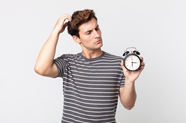 Hombre guapo joven que se siente desconcertado y confundido, rascándose la cabeza y sosteniendo un reloj despertador