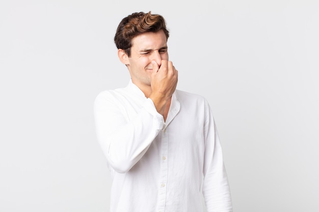 Hombre guapo joven que se siente disgustado, tapándose la nariz para evitar oler un hedor desagradable y desagradable