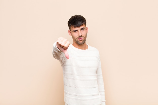 Hombre guapo joven que se siente enojado, enojado, molesto, decepcionado o disgustado, mostrando los pulgares hacia abajo con una mirada seria sobre la cálida pared