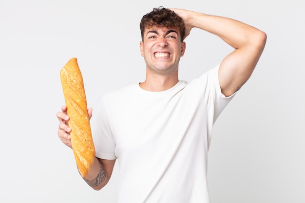 Hombre guapo joven que se siente estresado, ansioso o asustado, con las manos en la cabeza y sosteniendo una baguette de pan