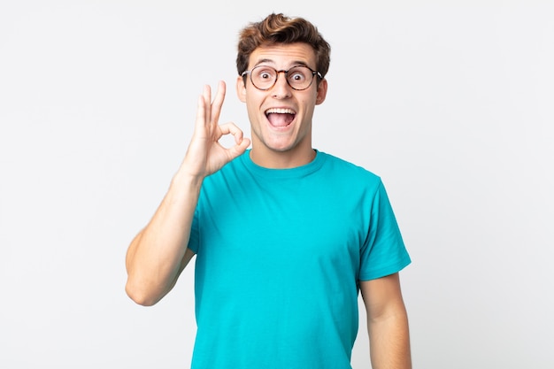 Hombre guapo joven que se siente exitoso y satisfecho, sonriendo con la boca abierta, haciendo un buen signo con la mano