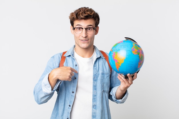 Hombre guapo joven que se siente feliz y apunta a sí mismo con un emocionado. estudiante sosteniendo un mapa del globo terráqueo