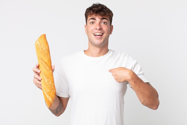 Hombre guapo joven que se siente feliz y apuntando a sí mismo con un emocionado y sosteniendo una baguette de pan