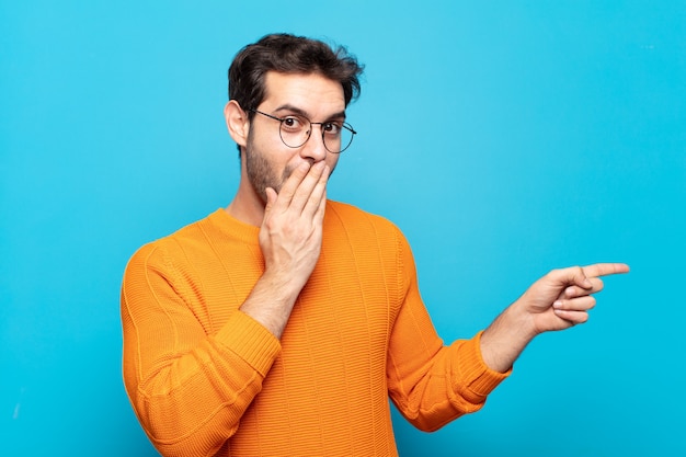 Hombre guapo joven que se siente feliz, conmocionado y sorprendido, cubriendo la boca con la mano y apuntando al espacio de copia lateral