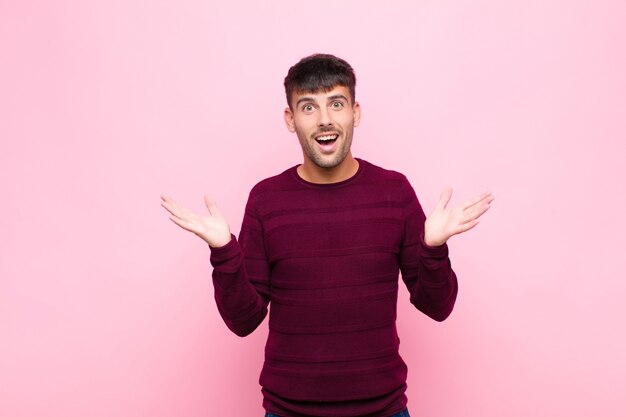 Hombre guapo joven que se siente feliz, emocionado, sorprendido o sorprendido, sonriendo y asombrado por algo increíble contra la pared rosa