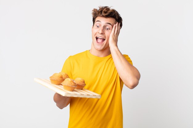 Hombre guapo joven que se siente feliz emocionado y sorprendido sosteniendo una bandeja de muffins