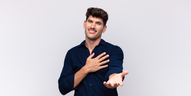 Hombre guapo joven que se siente feliz y enamorado, sonriendo con una mano al lado del corazón y la otra estirada al frente