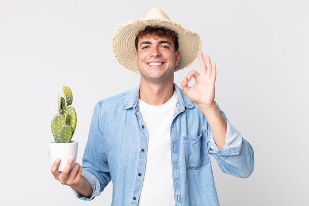 Hombre guapo joven que se siente feliz, mostrando aprobación con gesto bien. granjero sosteniendo un cactus decorativo