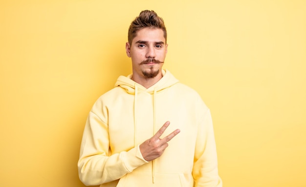 Hombre guapo joven que se siente feliz, positivo y exitoso, con la mano haciendo forma de v sobre el pecho, mostrando victoria o paz. extraño concepto de bigote