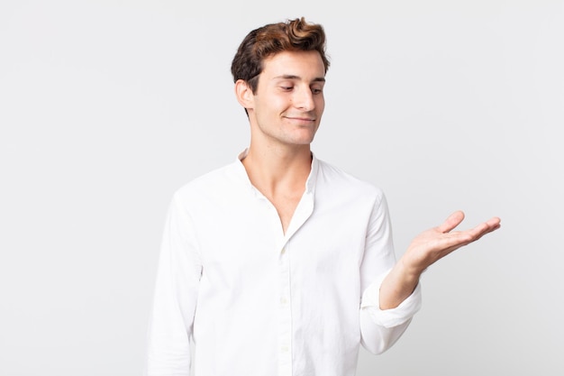 Hombre guapo joven que se siente feliz y sonriendo casualmente, mirando a un objeto o concepto sostenido en la mano al costado