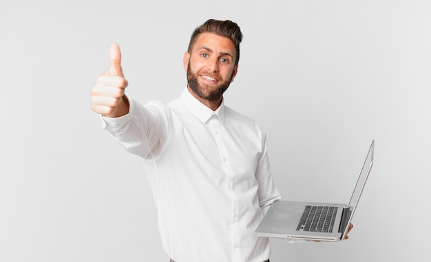 Hombre guapo joven que se siente orgulloso, sonriendo positivamente con los pulgares hacia arriba y sosteniendo una computadora portátil