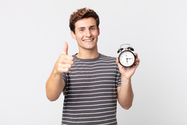 Hombre guapo joven que se siente orgulloso, sonriendo positivamente con los pulgares hacia arriba y sosteniendo un reloj despertador