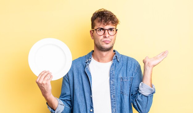 Hombre guapo joven que se siente perplejo, confundido y dudando. concepto de plato vacío