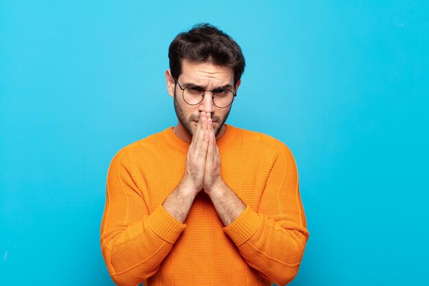 Hombre guapo joven que se siente preocupado, esperanzado y religioso, orando fielmente con las palmas presionadas, pidiendo perdón