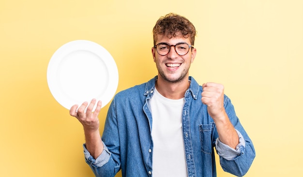 Hombre guapo joven que se siente sorprendido, riendo y celebrando el éxito. concepto de plato vacío