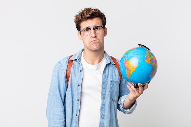 Hombre guapo joven que se siente triste y llorón con una mirada infeliz y llorando. estudiante sosteniendo un mapa del globo terráqueo