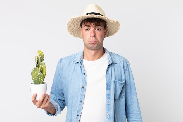 Hombre guapo joven que se siente triste y llorón con una mirada infeliz y llorando. granjero sosteniendo un cactus decorativo