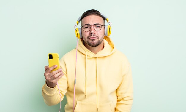 Hombre guapo joven que se siente triste y quejumbroso con una mirada infeliz y llorando. auriculares y concepto de teléfono inteligente