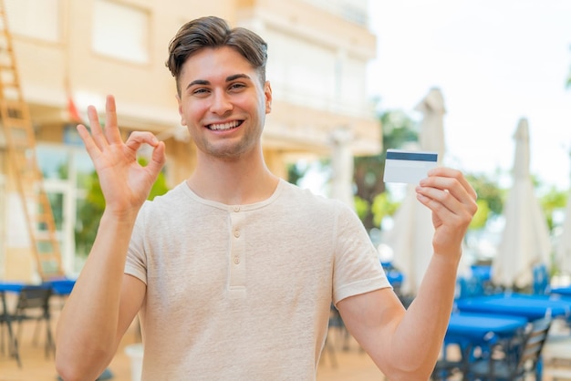 Hombre guapo joven que sostiene una tarjeta de crédito al aire libre que muestra el signo de ok con los dedos