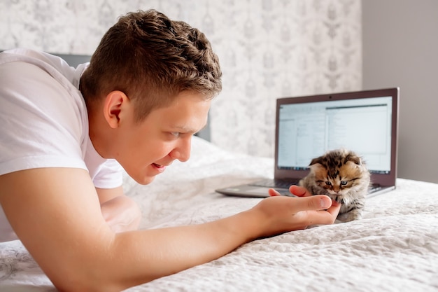 Hombre guapo joven que trabaja en casa mientras pone en cuarentena un coronovirus, acostado con una computadora portátil y un gatito en la cama