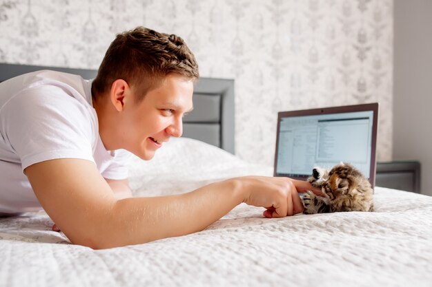 Hombre guapo joven que trabaja en casa mientras pone en cuarentena un coronovirus, acostado con una computadora portátil y un gatito en la cama